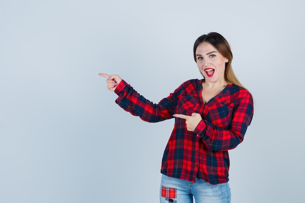 Young woman in checked shirt pointing left and looking happy , front view.