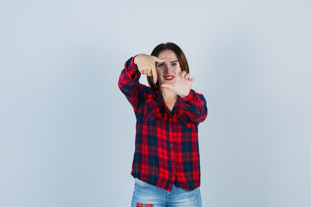 Foto giovane donna in camicia a quadri che mostra il gesto del telaio e sembra allegra, vista frontale.