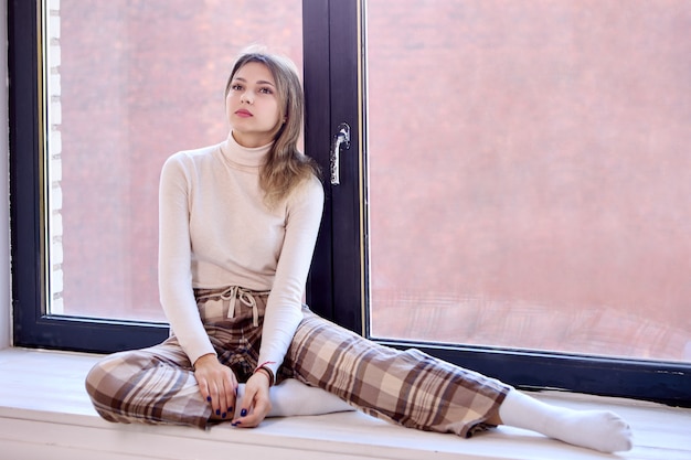 Young woman in checkered pants sits on windowsill