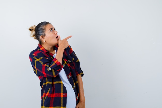 Young woman in checkered shirt pointing up, standing sideways and looking shocked .