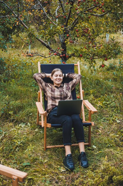 Young woman in a checkered shirt sitting in a chair outdoors in a garden and working on a laptop remote work female freelancer