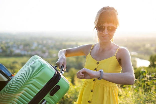 車のトランクにスーツケースを詰めながら時間をチェックする若い女性旅行と休暇のコンセプト