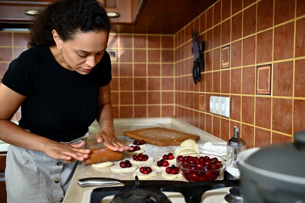 Young woman, chef pastry, rolls out dough with a rolling pin in round shapes on the kitchen countertop, prepares homemade delicious cherry dumplings