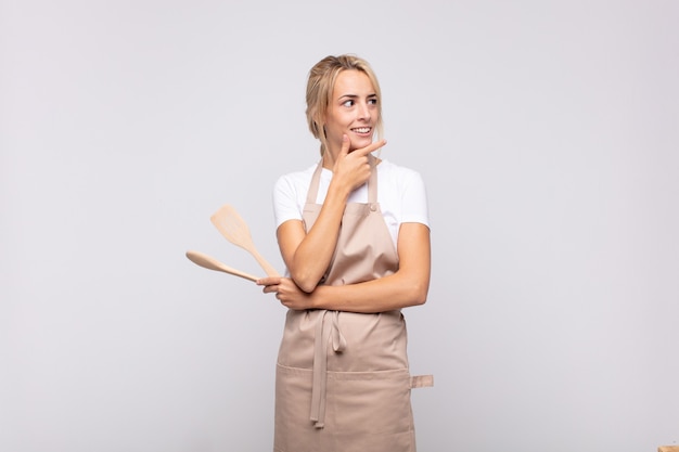 Young woman chef smiling with a happy, confident expression with hand on chin, wondering and looking to the side