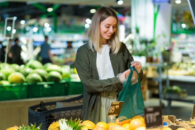 若い女性はスーパーマーケットでエコバッグ野菜や果物オレンジを選んで選びます。カウンターの近くに食料品店を立っている女性客が、市場で再利用可能なパッケージを購入して投入します