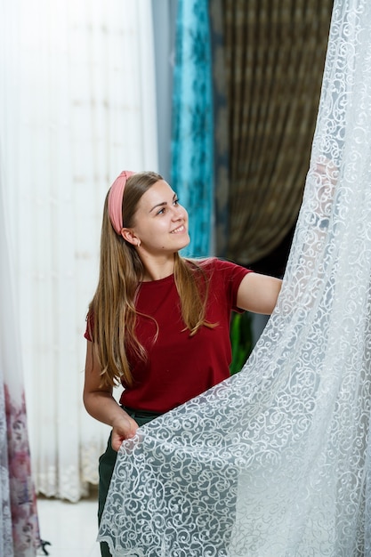 Giovane donna che sceglie tessuto per nuove tende in un negozio. i campioni della tenda sono appesi ai ganci su un binario nel negozio. campioni di trame di tessuto, tulle e tappezzeria di mobili.