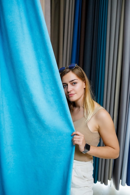 店で新しいカーテンの生地を選ぶ若い女性。カーテンのサンプルは、店内のレールのハンガーに掛けられています。生地、チュール、家具の装飾のテクスチャのサンプル。