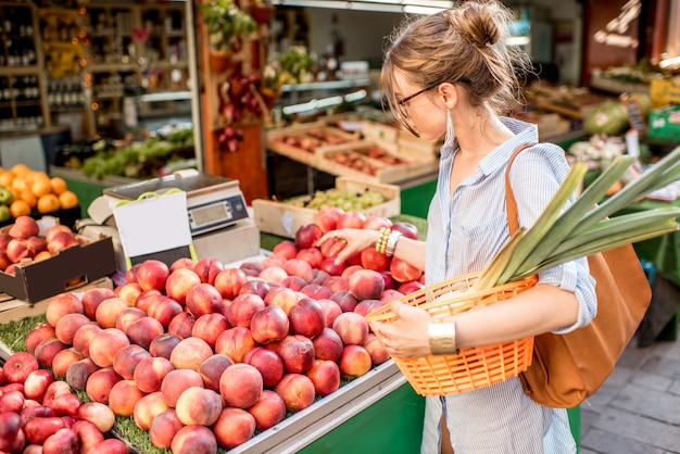 フランスの食品市場でバスケットと立っている新鮮な桃を選ぶ若い女性