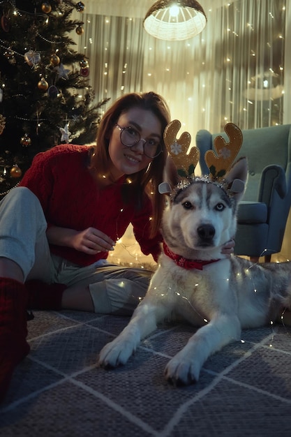 Молодая женщина в рождественский вечер вместе с собакой весело надевает рождественские оленьи рога и гирлянды, сидя на полу перед рождественской елкой, смотрит в камеру