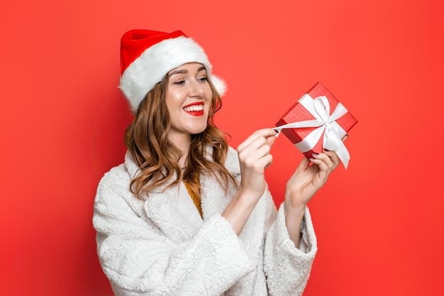 Young woman in christmas holiday hat smiling and opening red gift box  isolated over red background