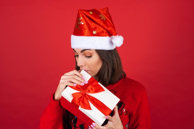 Молодая женщина в рождественском свитере и новогодней шапке с подарочной коробкой заглядывает внутрь