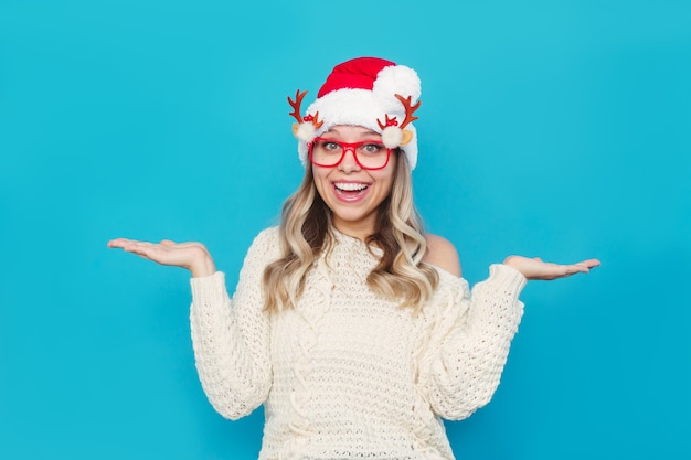 크리스마스 흰색 니트 스웨터와 산타클로스 모자를 쓴 젊은 여성이 의심스러운 표정으로 손을 펼치고 있다