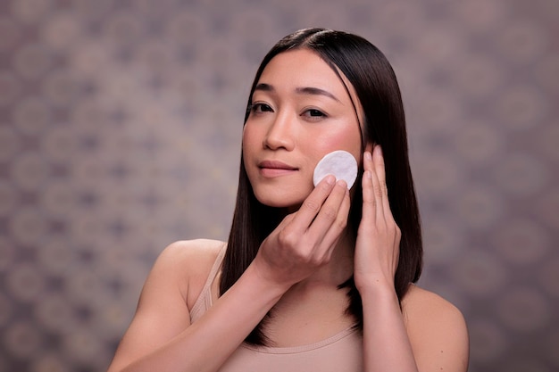 ミセル水で綿のディスクを使用して顔を掃除する若い女性。装飾的な化粧品から顔を洗い、新鮮な輝く肌をクレンジングし、スキンケア治療を行う魅力的なアジアの女性