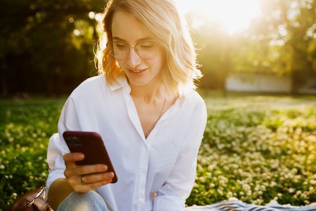 Il primo piano della giovane donna in camicia bianca legge il messaggio al telefono cellulare nel parco.