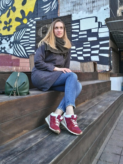 Молодая женщина в пальто, джинсах и кроссовках сидит на деревянных ступенях окрашенной стены
