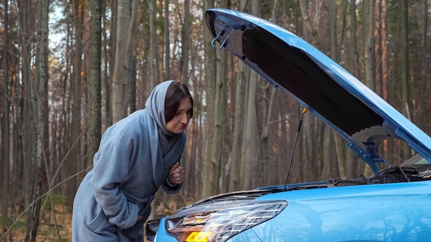 Young woman in coat and scarf on head looks under hood of broken car with alarm headlights on roadside near autumn forest with bare trees