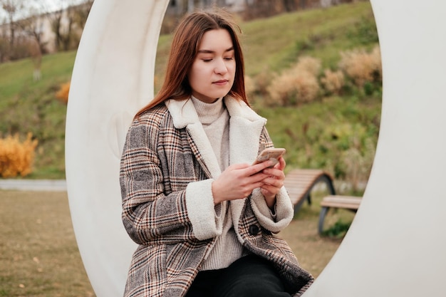 Young woman in coat sitting on white swing with smartphone in hands in autumn park Working on mobile device out of home outdoors Female freelancer blogger Texting chatting reading