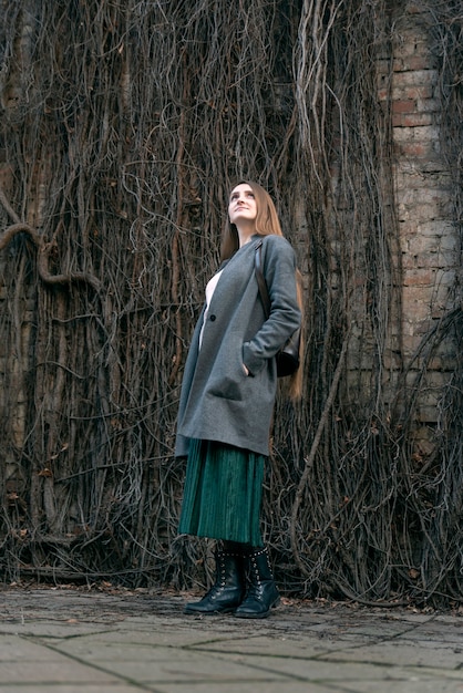 Young woman in a coat stands against the backdrop of a wall of withered ivy and looks up. Side view. Vertical frame