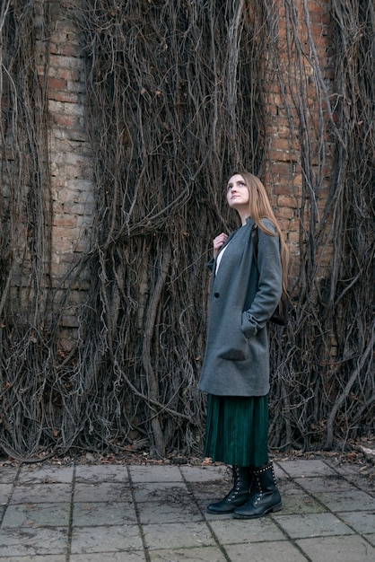 Young woman in a coat stands against the backdrop of a wall of withered ivy. Side view. Vertical frame.