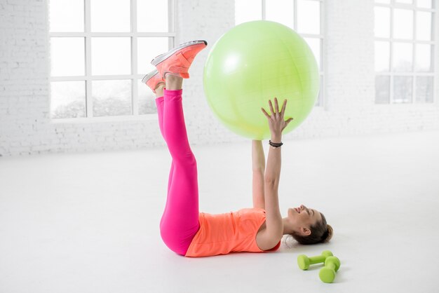Молодая женщина в красочной спортивной одежде растягивается с фитнес-мячом в белом тренажерном зале