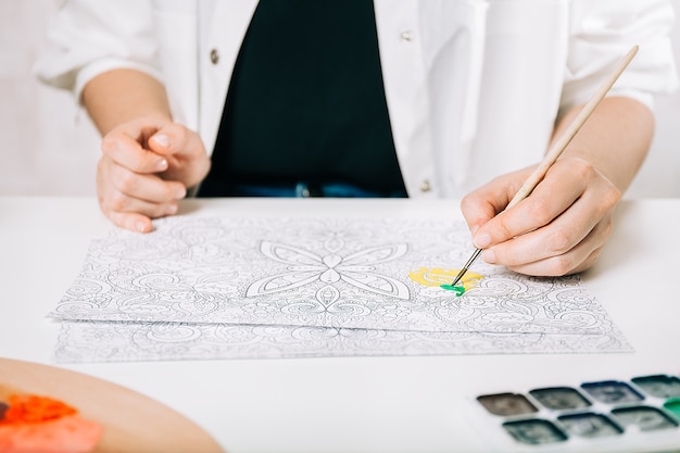 Young woman coloring page antistress at table indoors, mental wellbeing and art therapy. Woman paints a sketch, meditative process of coloring pages