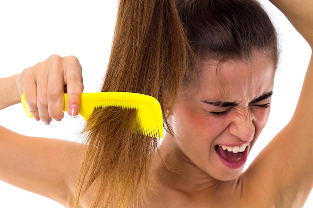 Young woman combing her long brown ponytail using yellow hair brush on white background in studio