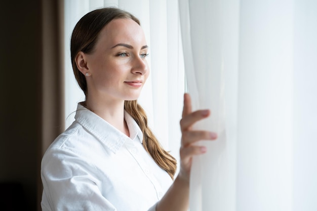 young woman comes to bright window opening white curtain to enjoy city view satisfied female guest