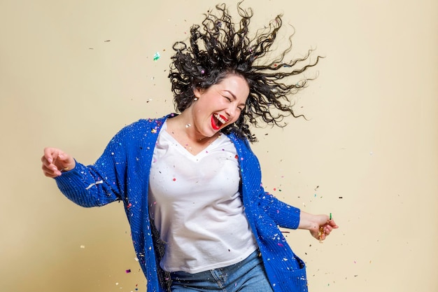 A young woman in confetti laughs and dances Beautiful emotional curly brunette in a white Tshirt blue sweater and jeans Holidays and events Closeup