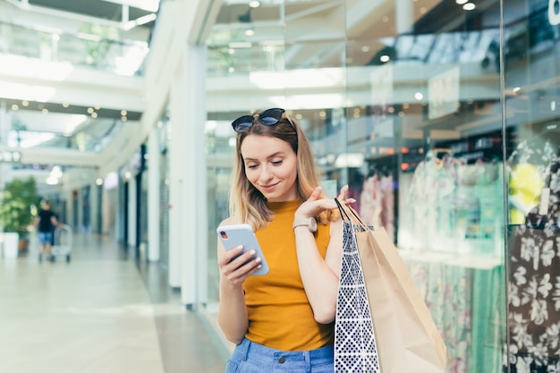 モール内の若い女性の消費者は、スマートフォンを使用してチャットを閲覧し、使用しています。ショッピングセンターで彼女の手で携帯電話を持って立っている女性。屋内。ギフトバッグを持った幸せな買い物客の女の子が購入する