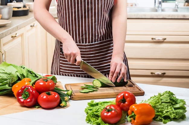 自宅のキッチンで料理をする若い女性。健康食品。ダイエット。ダイエットの概念。健康的な生活様式。家で料理。食べ物を用意します。女性がきゅうりと野菜をナイフで切る。