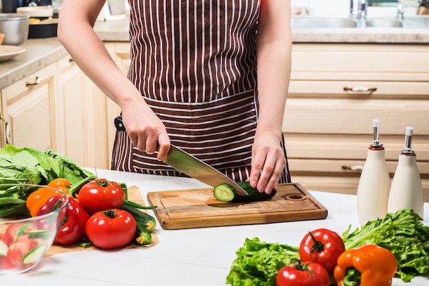 Giovane donna che cucina in cucina a casa. cibo salutare. dieta. concetto di dieta. uno stile di vita sano. cucinare a casa. prepara da mangiare. una donna taglia un cetriolo e delle verdure con un coltello.