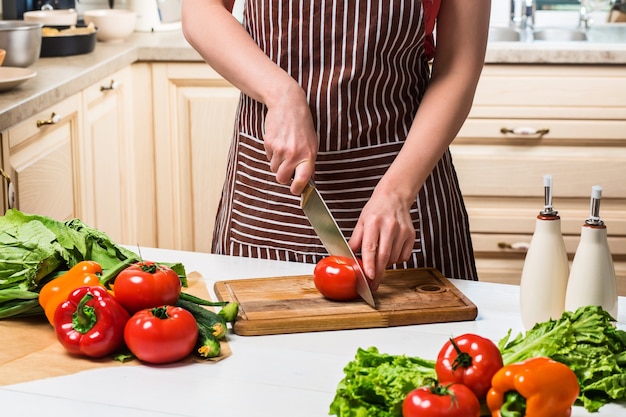 自宅のキッチンで料理をする若い女性。健康食品。ダイエット。ダイエットの概念。健康的な生活様式。家で料理。食べ物を用意します。女性がトマトと野菜をナイフで切る。