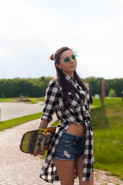 Young woman in cool glasses and short briefs is holding her skate behind on the background of park.