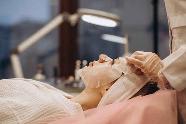 Молодая женщина-косметолог наносит маску из белой глины на лицо женщины Красивая брюнетка в салоне красоты
