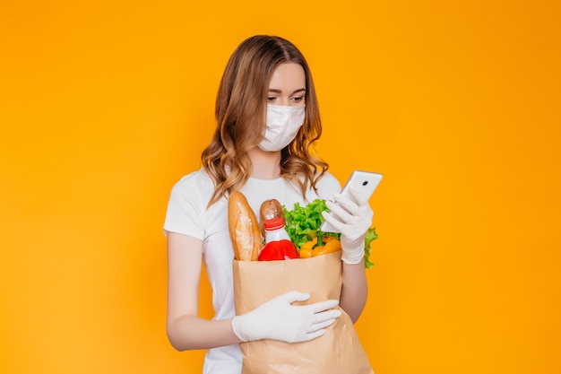 Young woman courier wearing a medical mask looks at a mobile phone and holds an eco paper bag with food