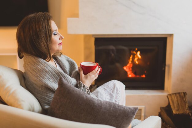 The young woman covered with a cozy warm blanket holding a cup of hot drink Woman sitting with her eyes closed on the background of the fireplace