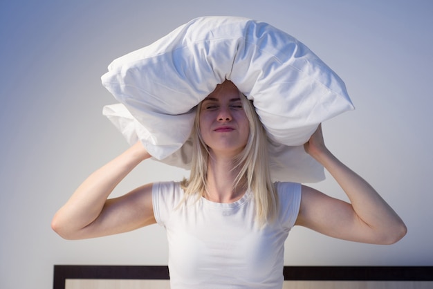 Young woman covering ears with pillow because of noise.