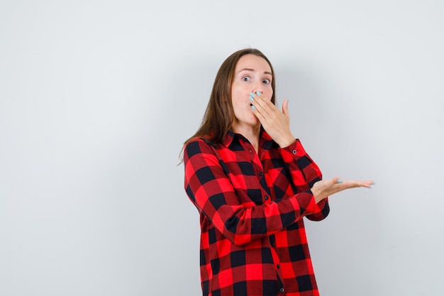 Photo young woman covering mouth with hand, stretching hand as holding something in checked shirt and looking shocked. front view.