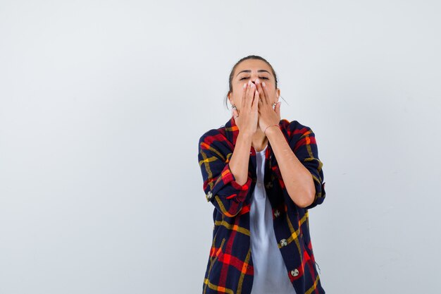 Young woman covering mouth with hands in checkered shirt and looking curious , front view.