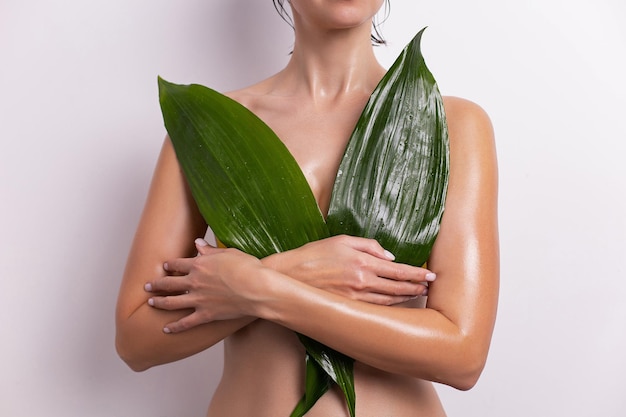 young woman covers her breasts with a green leaf of a tropical plant in summer on a white background