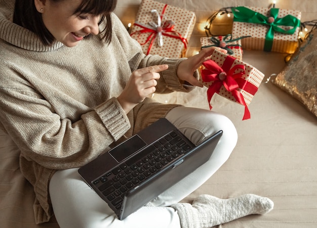 A young woman in a cozy sweater buys Christmas presents on the Internet. Concept of choosing gifts online and distance giving.