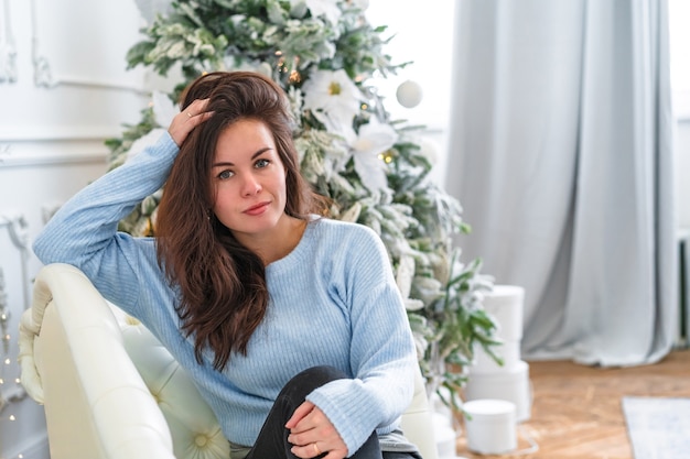 A young woman in a cozy sweater sits on a sofa in front of a decorated Christmas tree