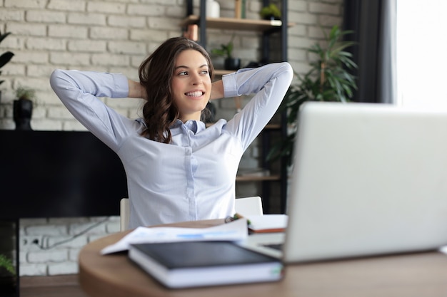 Young woman crossed hands behind head, enjoying break time at home. Peaceful carefree business woman resting at table with computer, looking aside, dreaming of future.