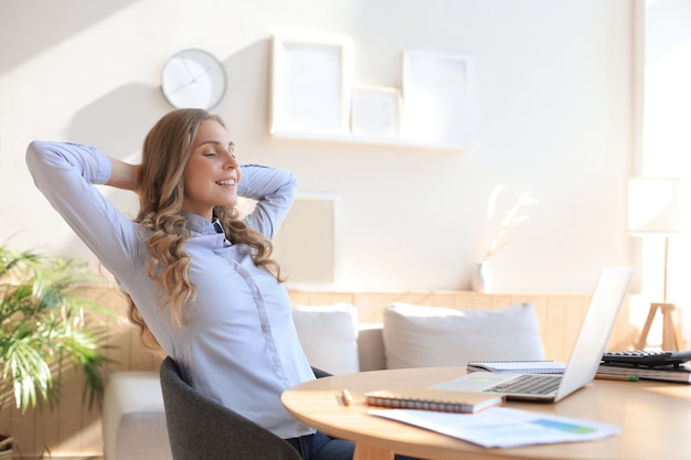 young woman crossed hands behind head, enjoying break time at home. peaceful carefree business woman resting at table with computer, looking aside, dreaming of future.