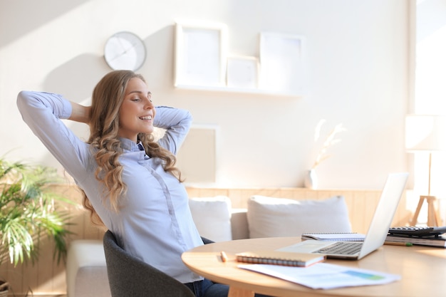 Young woman crossed hands behind head, enjoying break time at home. Peaceful carefree business woman resting at table with computer, looking aside, dreaming of future.