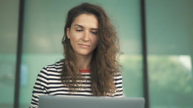 Young woman curly hair with laptop in stripped sweater glass office window