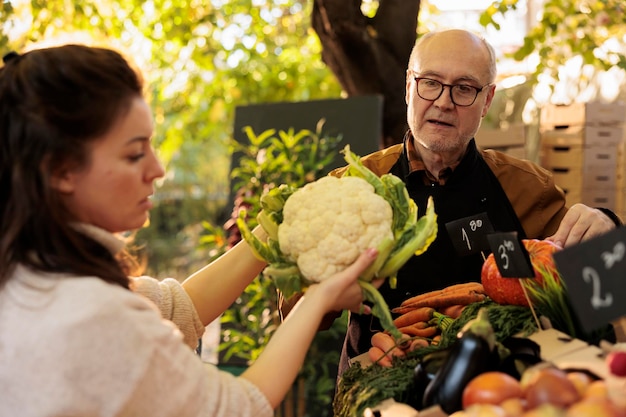 Photo young woman customer holding cauliflower while buying fresh locally grown organic vegetables at local farmers market. mature male vendor serving female consumer while selling his home-grown produce