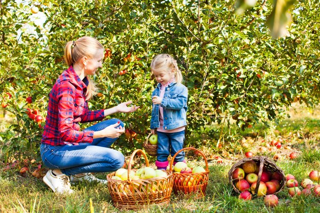 Young woman and a cute small girl at the apple orchard with green trees and grass on a background