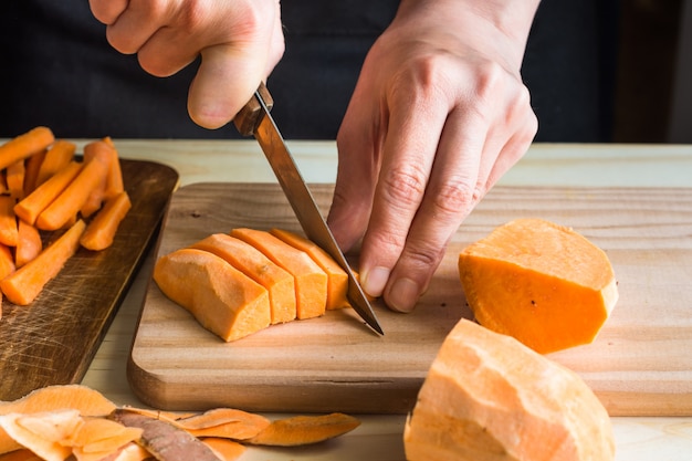 Young woman cutting with knife sweet potato into wedges peels on wood table