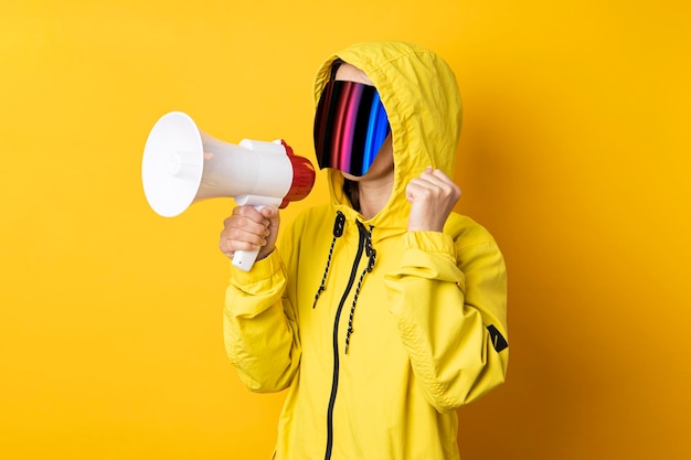 Young woman in cyberpunk glasses with a megaphone in a yellow\
jacket on a yellow background